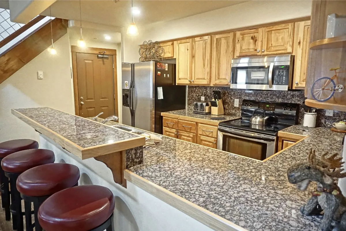A modern kitchen with granite countertops, wooden cabinetry, stainless steel appliances, and bar stools lining a breakfast bar. The setup invites conversation and shared meals, while decorative accents add warmth and personality.