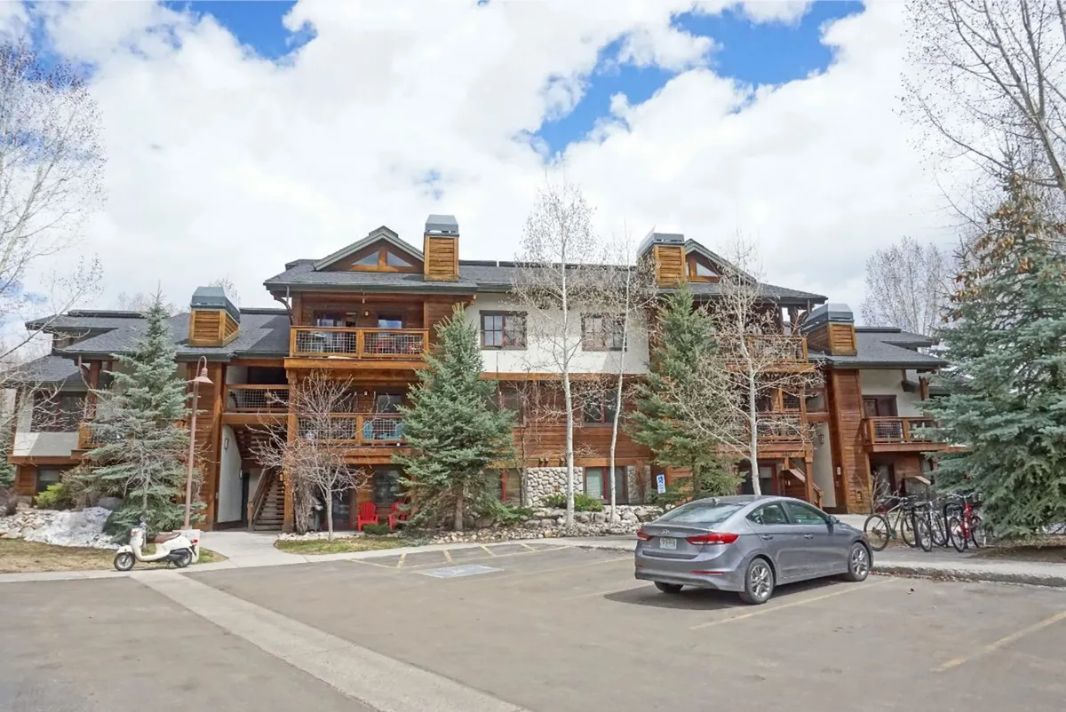 This exterior image features the building's wooden architecture surrounded by mature trees and a visible parking area. Balconies overlook the natural setting, suggesting an easy transition from indoors to the outdoors. The atmosphere feels welcoming and is set against a backdrop of greenery, hinting at close proximity to parks, trails, and family-friendly activities.