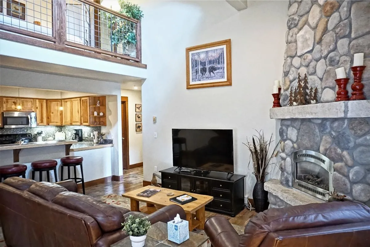 A spacious, open-concept living area featuring a striking stone fireplace, plush leather sofa, wooden coffee table, television, and a bar area with stools. Large windows let in natural light and hint at mountain views, and the space feels connected, warm, and inviting.