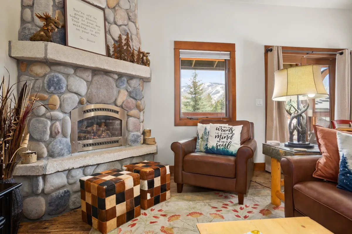 The living space features a stone fireplace, comfortable seating with colorful pillows and ottomans, and a relaxed, social ambiance. Large windows suggest mountain views, enhancing the sense of place and leisure.