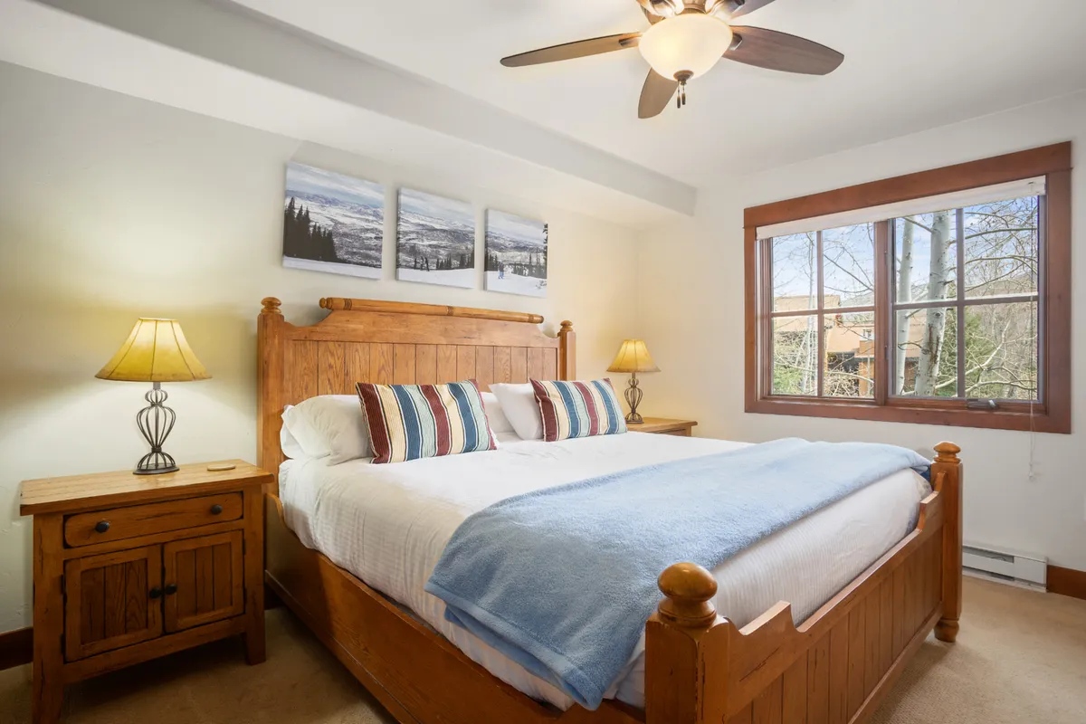 A serene bedroom with a wooden bed frame, striped pillows, decorative wall art, and soft lighting from bedside lamps. Natural light fills the room, suggesting a peaceful retreat for rest and relaxation.