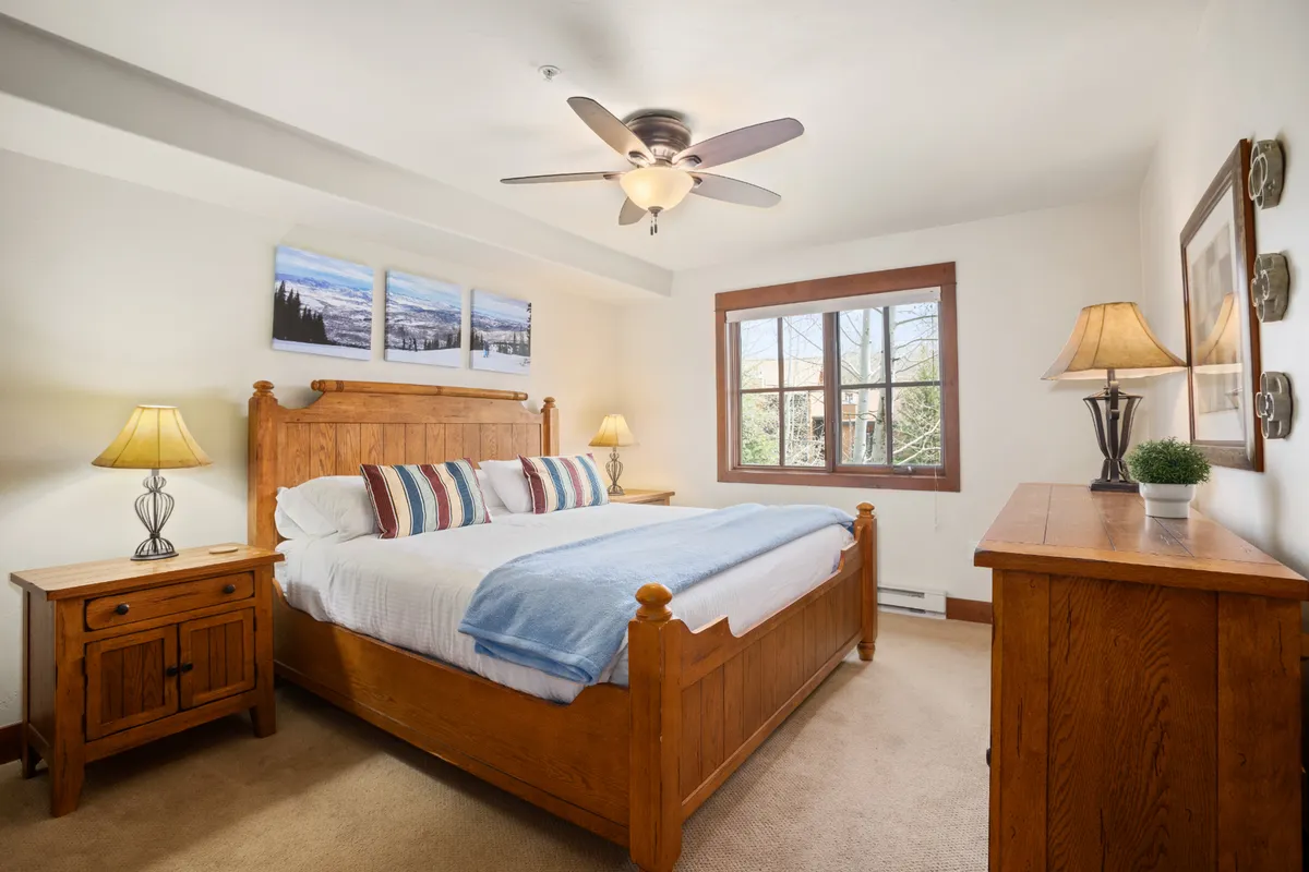 A calm and restful bedroom with a wooden bed, striped pillows, nightstand with lamp, and a window offering serene mountain views. The space feels cozy and inviting, ideal for unwinding after a day of adventure.