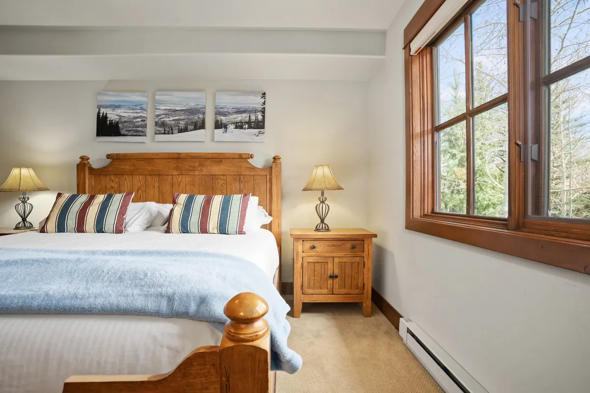 A cozy bedroom scene with a bed adorned with striped pillows, a wooden bedside table, and warm lamp lighting. The atmosphere is inviting and restful, ideal for relaxing or reading after a day exploring with friends.