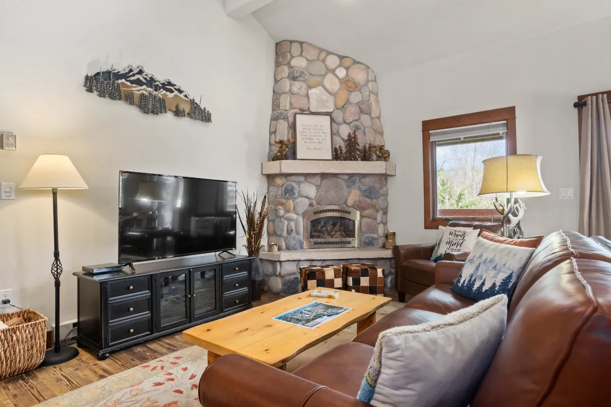 A bright, welcoming living room featuring a stone fireplace, leather sofa, coffee table, and TV. Sunlight streams in through large windows, accentuating the soaring ceiling and creating a sense of open, airy space. The seating area is arranged for both relaxation and social gatherings, offering a cozy yet functional environment.