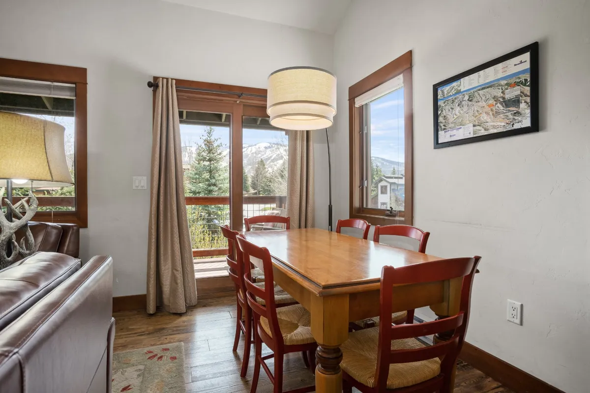 A bright dining area with a wooden dining table, elegant light fixture, and large windows framing scenic views. The setup encourages shared meals, game nights, and relaxed gatherings, with natural light streaming in.