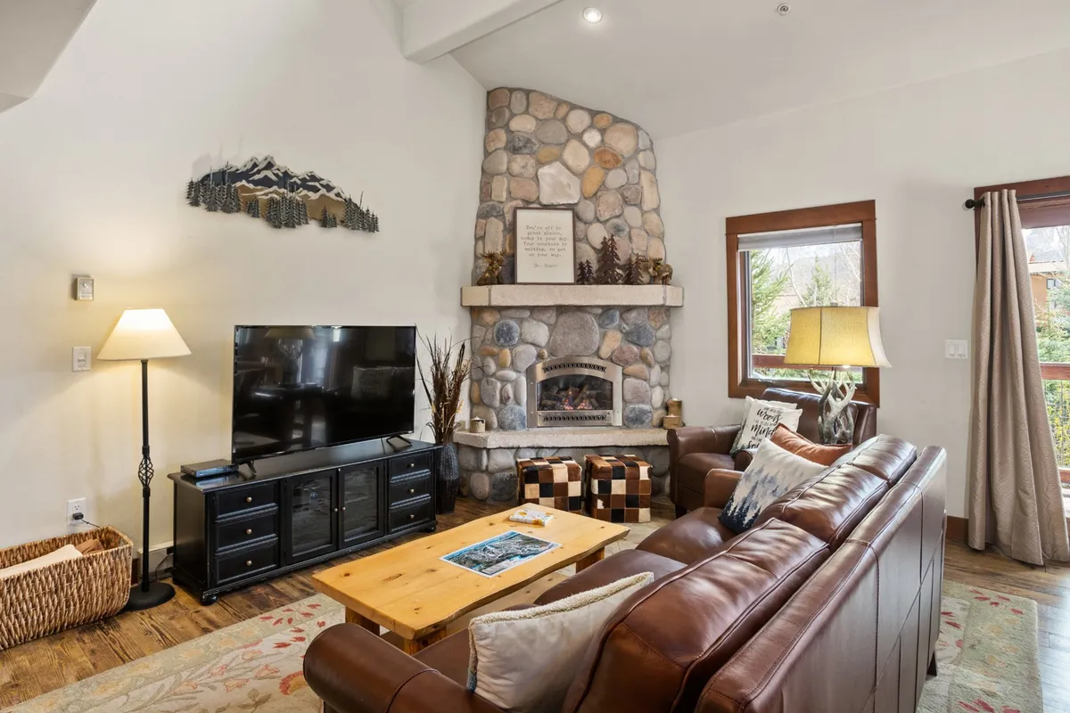 A cozy living area anchored by a stone fireplace, leather sofa, and wooden coffee table. The room is warm and inviting, with decorative wall art and a TV, evoking a mountain lodge atmosphere perfect for relaxing after a day on the slopes.