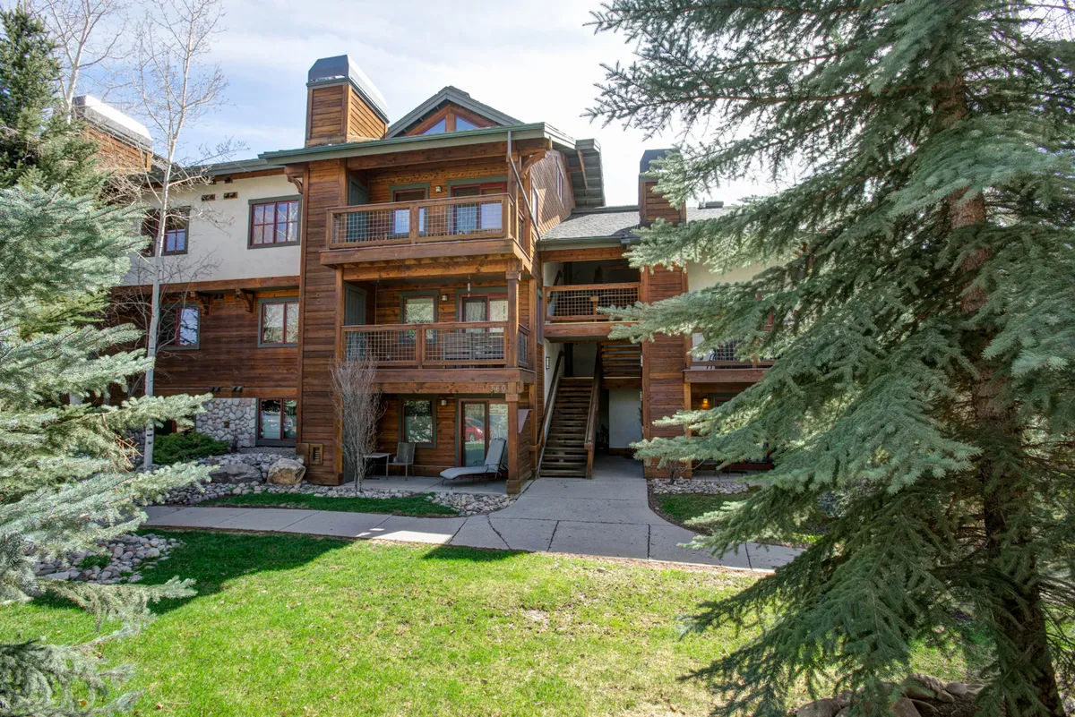The hero image showcases the condo's inviting wooden balcony overlooking a lush green lawn, with a charming stone pathway winding through vibrant gardens. The outdoor space is perfect for both peaceful mornings and social evenings, set against the backdrop of Steamboat Springs' natural beauty.