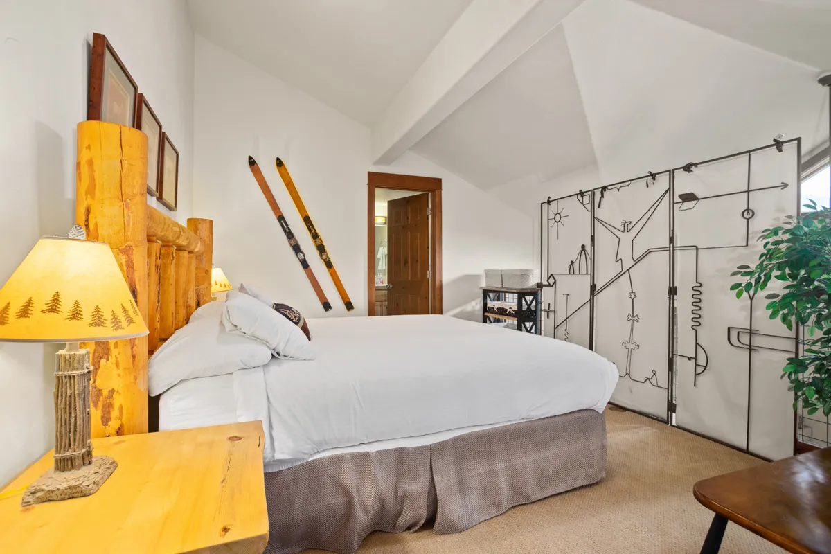 A tranquil loft space featuring a rustic log bed, decorative ski poles, soft lighting from lamps, a screen divider for privacy, and potted plant accents. The décor blends comfort with mountain charm, emphasizing flexible and semi-private sleeping arrangements.