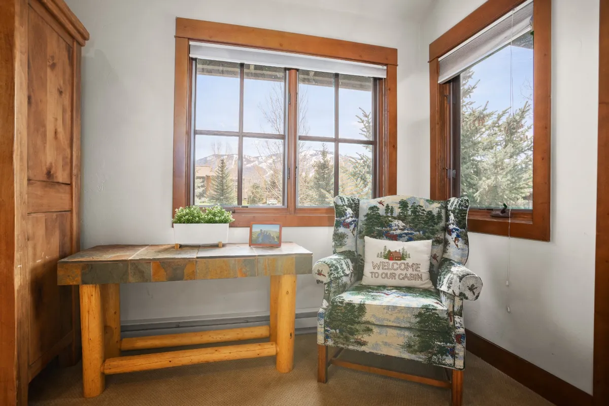 This interior image captures a wooden table and a cozy armchair with a forest pattern positioned beside large windows that offer views of the outdoors. The generous natural light and visible greenery outside reinforce the connection to nature and a sense of relaxation after outdoor activities.