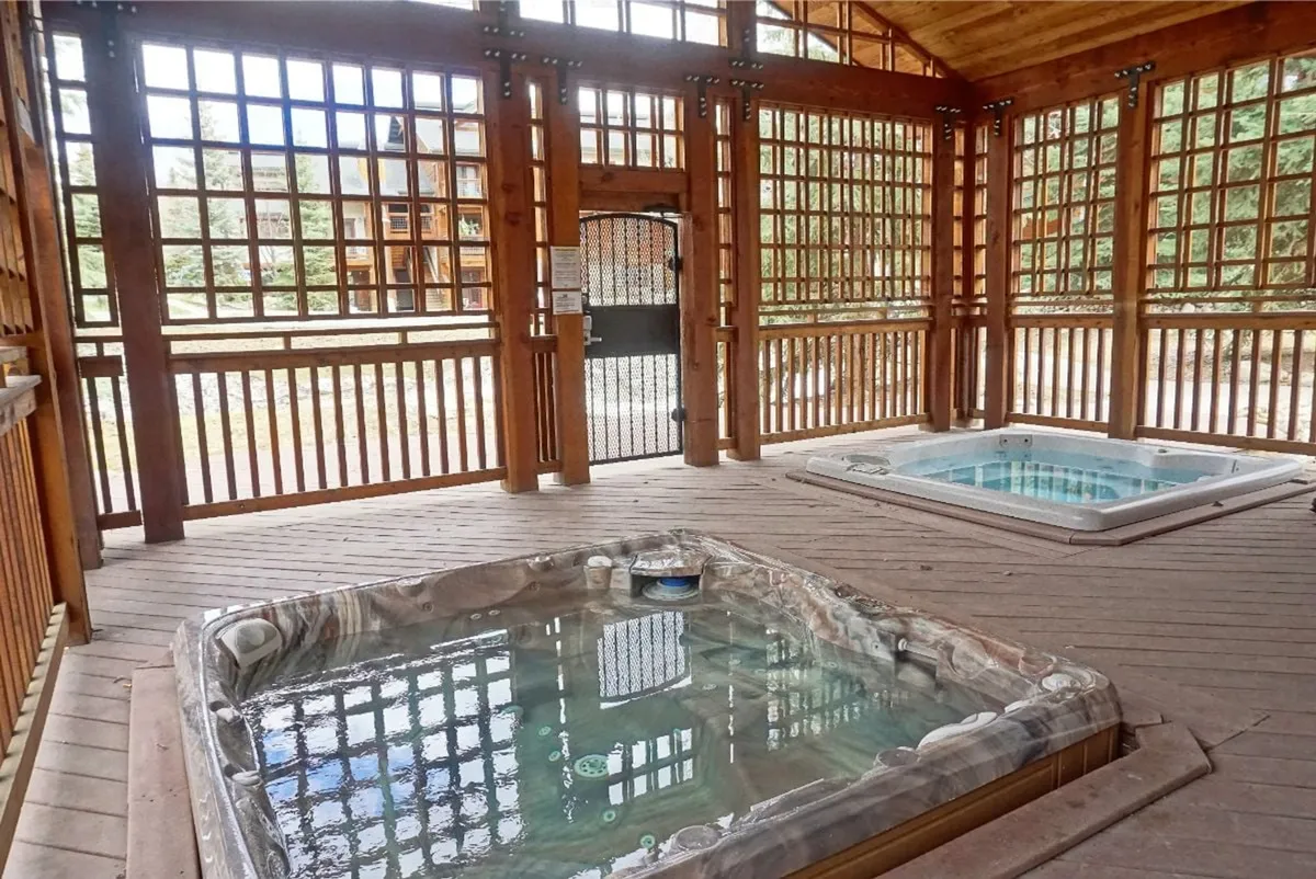 This image features two outdoor hot tubs nestled beneath a wooden lattice pergola, with an open doorway leading outdoors. The scene is serene, with the hot tubs invitingly steaming, suggesting relaxation in all seasons. The hot tubs' proximity to the outdoors under a protective cover matches the description in the section.